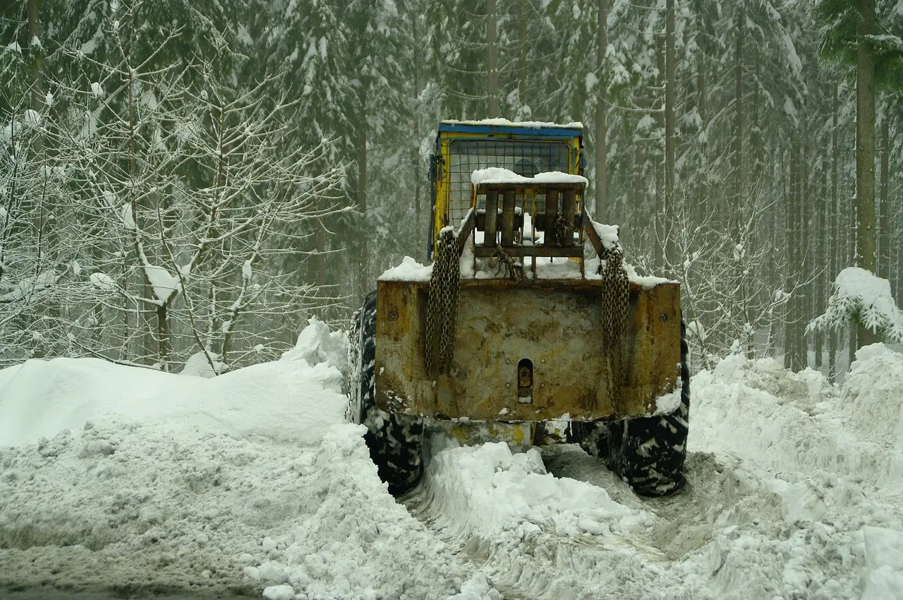 A large yellow and blue snowplow vehicle with weathered paint and accumulated snow works diligently, pushing through deep snow to clear a path in a quiet, snow-covered forest under a soft winter light.