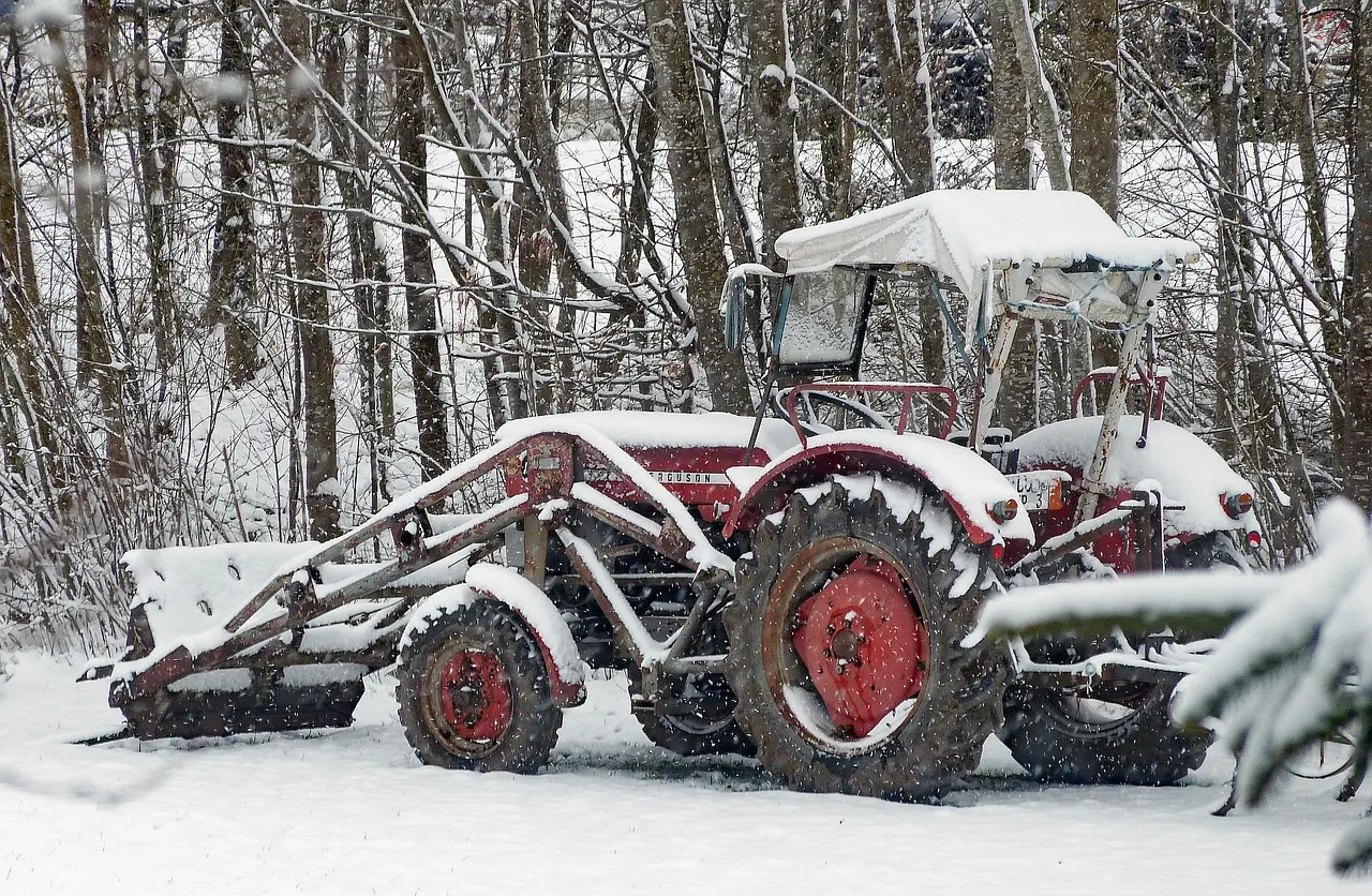 A red tractor with the marking "ACUSON D 3" sits stationary, covered in a thick layer of fresh snow, equipped with a front-mounted snow-clearing blade, amidst a peaceful snowy landscape of frosted trees.