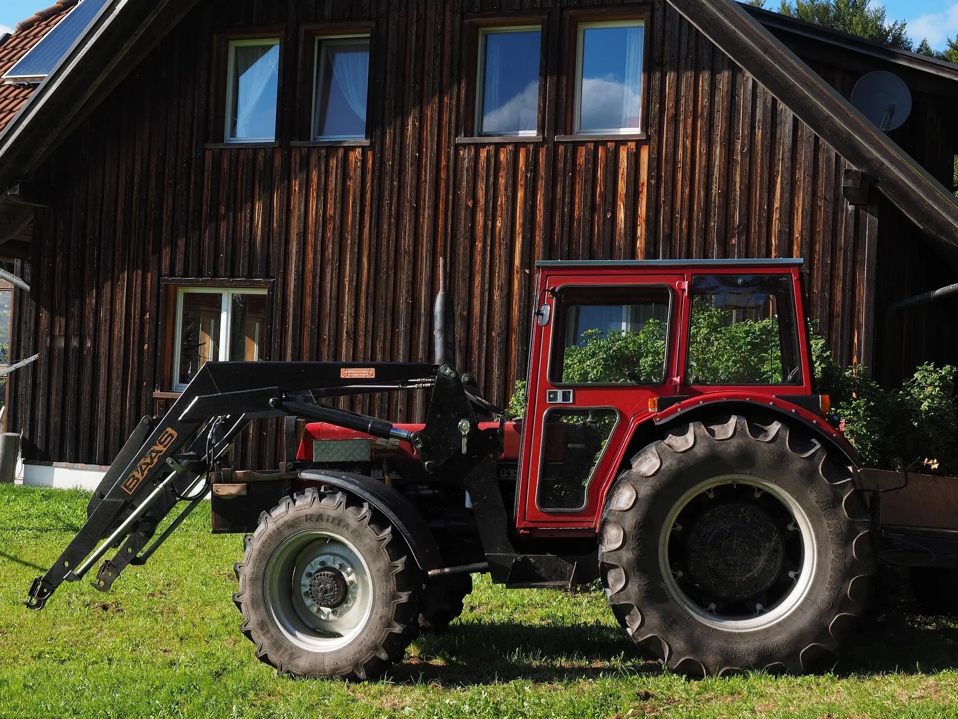 A red utility tractor with a black front loader parked in front of a rustic wooden cabin. The cabin has solar panels on its roof, and text on the image reads "BAAS RALLYE R".