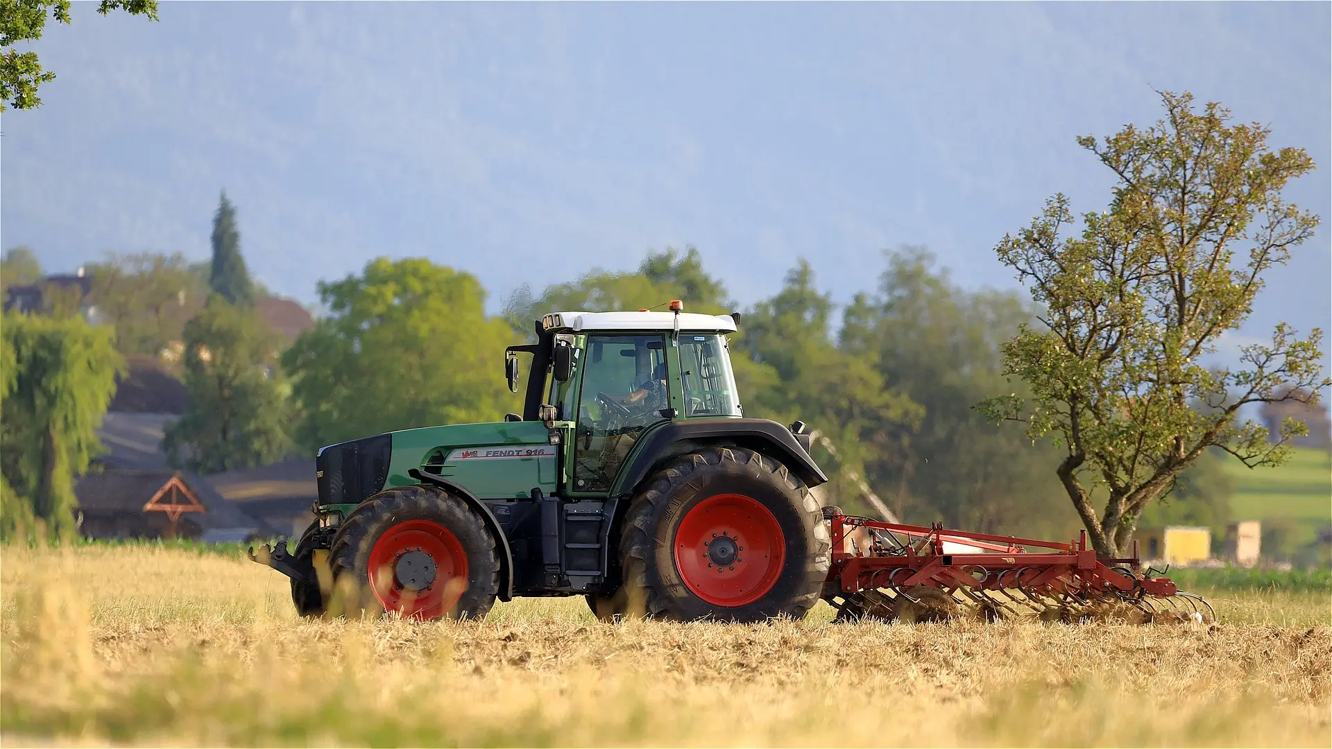 A Fendt 916 tractor in green with red wheels plows a vast, brown field. Farm buildings and trees are visible in the background under a hazy sky.