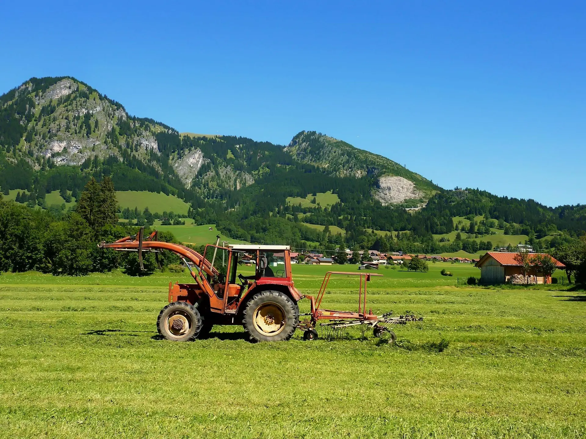 A red tractor with a hay tedder works in a lush green meadow, with mountains and a small village visible in the distance under a clear blue sky.