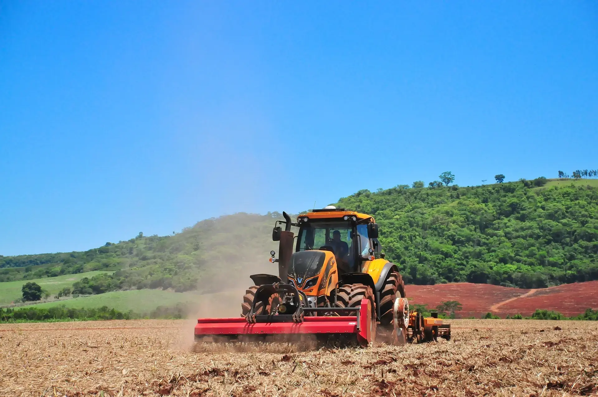 A yellow and black tractor with red attachments tills a dry, brown field, kicking up dust under a clear blue sky with green hills in the distance.