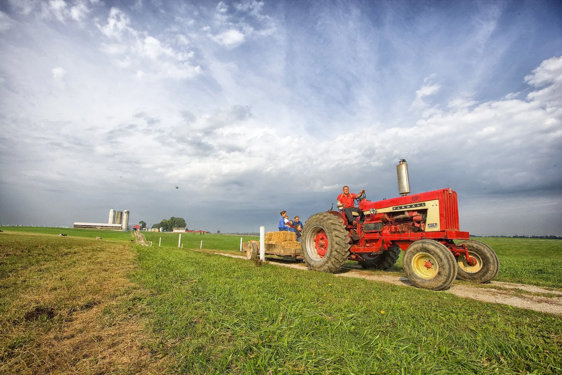 A person in a red shirt drives a vibrant red vintage Farmall tractor with yellow-rimmed wheels along a dirt path in a lush green field. Behind, a wooden trailer loaded with hay bales carries several passengers. Farm buildings, including white silos, are visible in the background under a partly cloudy sky. Text on the tractor reads "FARMALL" and "BOC." The scene evokes a serene moment of rural life.