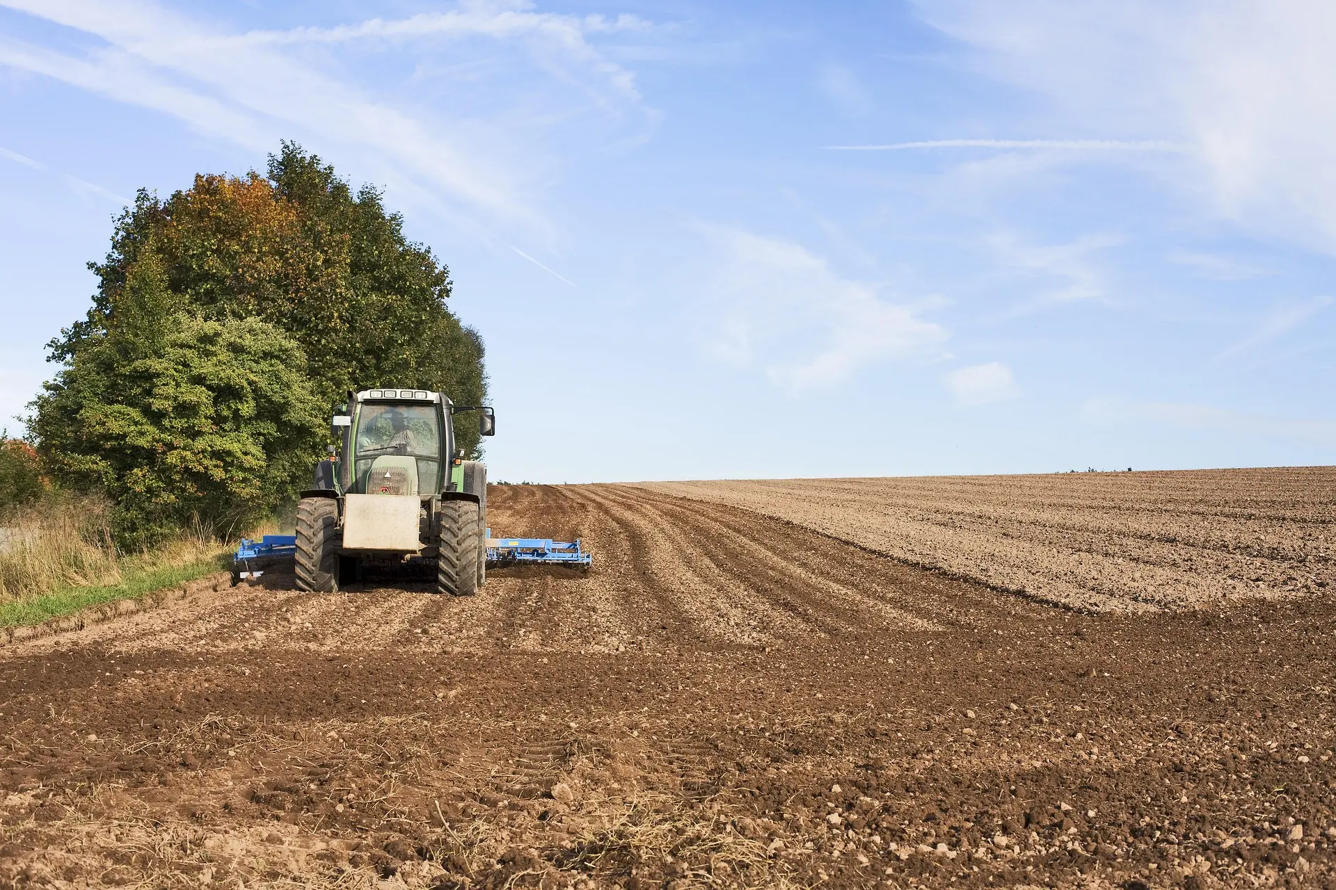 A green tractor with a blue tillage implement creates neat furrows in a dark brown field. A line of green and yellow trees stands on the left under a bright blue sky with white clouds.