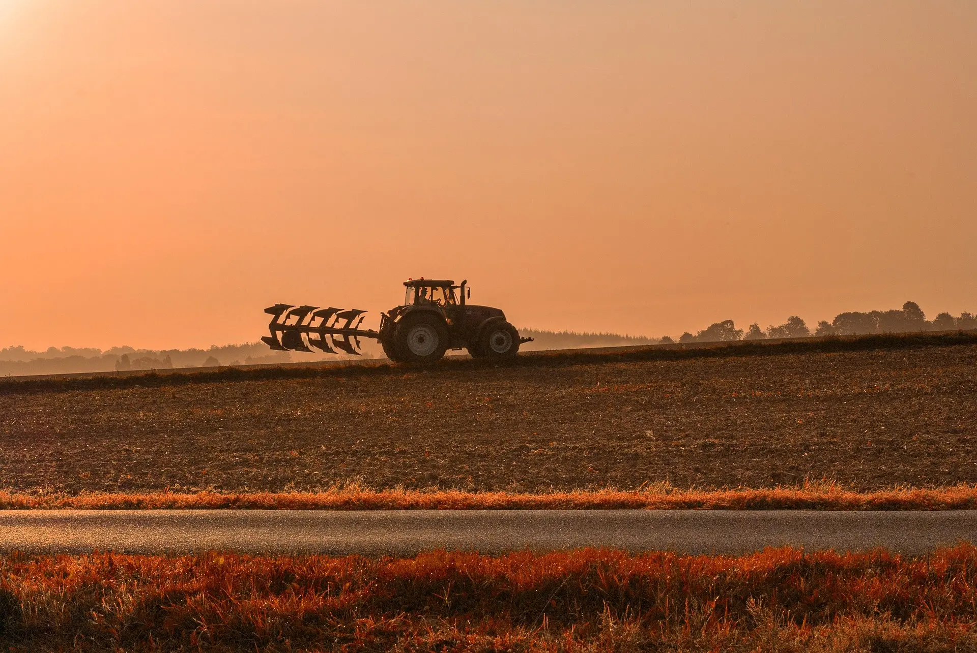 A tractor pulling a multi-furrow plow works along a straight rural path at sunset or sunrise, with the warm glow of orange and yellow light coloring the sky and fields, and blurred tree lines in the background.