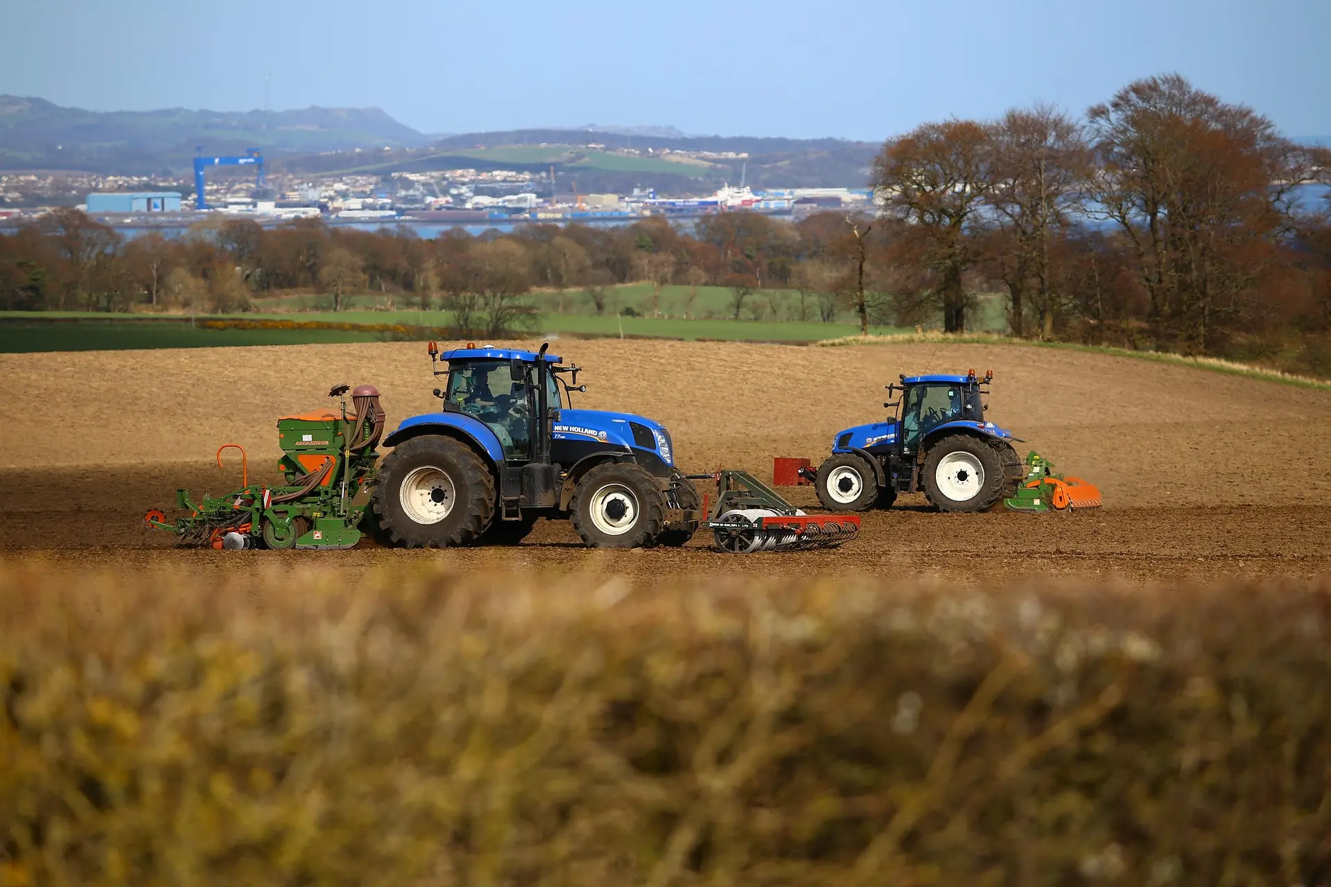 Two blue tractors equipped with different modern farming implements, including a seed drill and a plow, work in a field. One features a GPS navigation system for precision agriculture, with industrial buildings visible in the distance under a light blue sky.