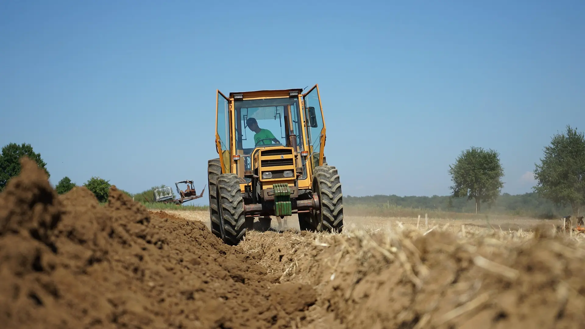 A yellow tractor with an open door drives across freshly tilled soil, creating two neat furrows. The driver wears a green shirt and is focused on operating the machine. In the background are harvested fields with remaining stubble, a few distant trees, and a clear blue sky.