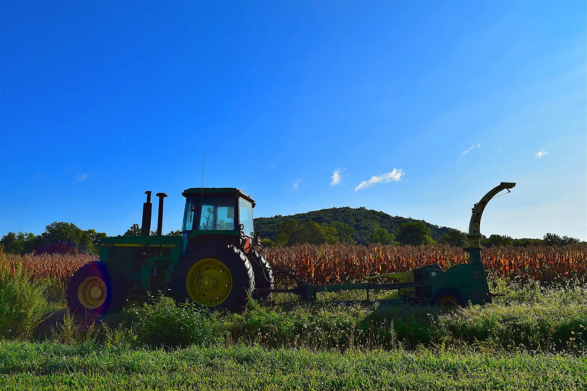A green John Deere tractor with large rear tires featuring yellow hubs is parked in a field of red crops, connected to an agricultural implement with a curved top section. Green weeds are visible in the foreground, with a distant green hill and a blue sky with white clouds completing the rural scene.