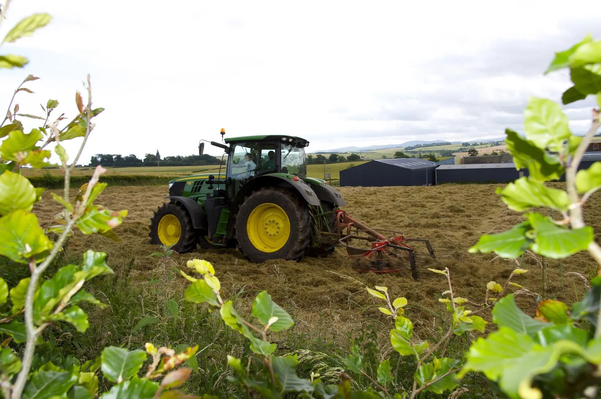 A green and yellow tractor actively pulling a red agricultural tiller, creating rows in a field of golden hay stubble. Green plants are in the foreground under a cloudy sky, with farm buildings and trees in the distance.