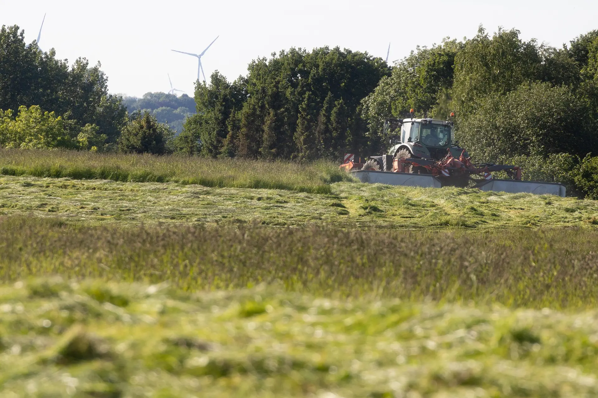 A white and red tractor carrying agricultural machinery cuts grass in a lush green field, showcasing modern farming. The scene, under a bright sky, includes dense trees and distant wind turbine towers, highlighting the integration of agriculture and sustainable energy.