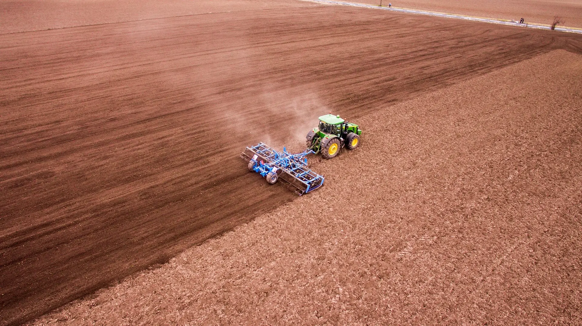 An aerial photograph showcasing a green tractor and attached blue equipment methodically tilling a vast, brown field. The scene highlights the neat, geometric patterns left in the soil, with distant field borders and figures providing scale, emphasizing the precision of mechanized agriculture.