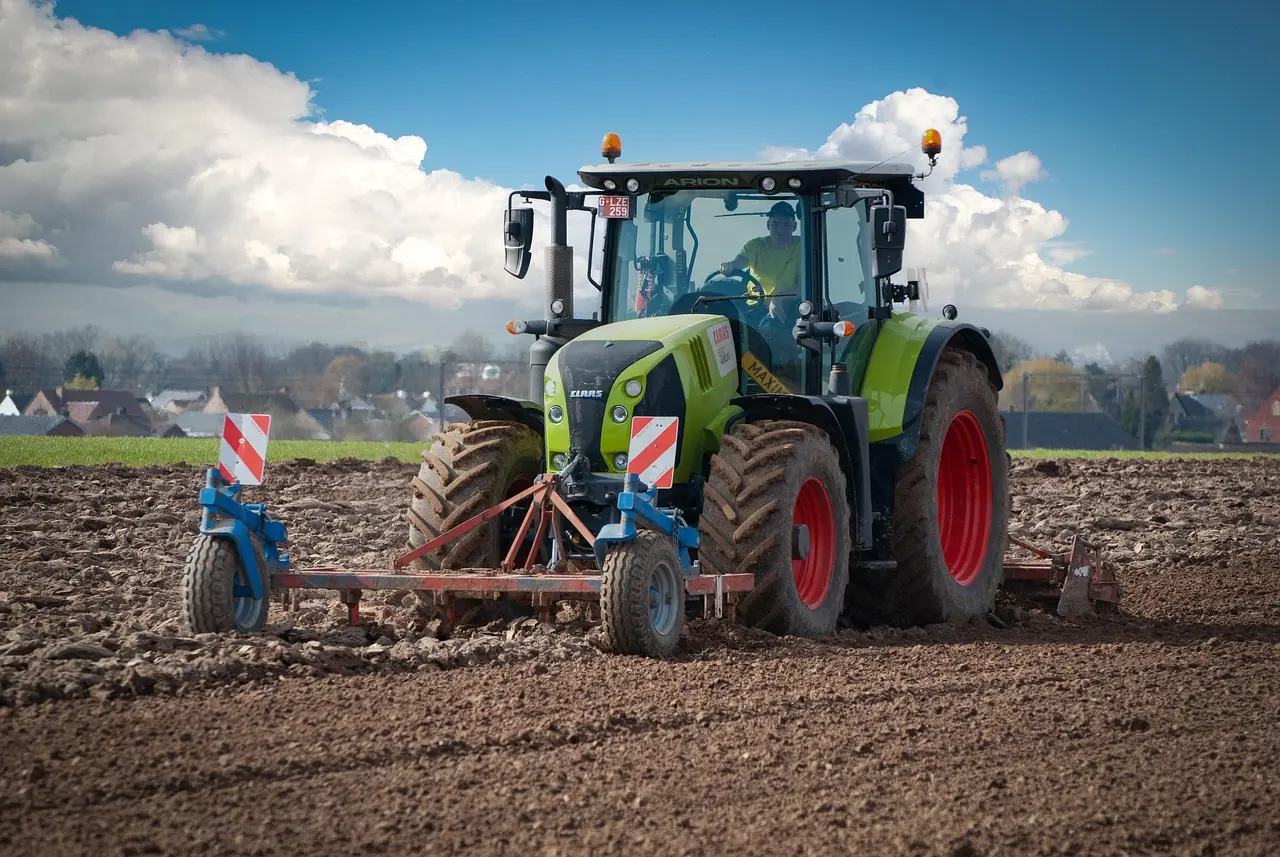 A vibrant green CLAAS ARION tractor, featuring prominent orange warning lights, actively pulls a blue tillage implement through rich brown soil. The sharp focus on the tractor and its detailed branding contrasts with the soft background of a blue sky, distant houses, and trees, highlighting modern agricultural machinery at work.