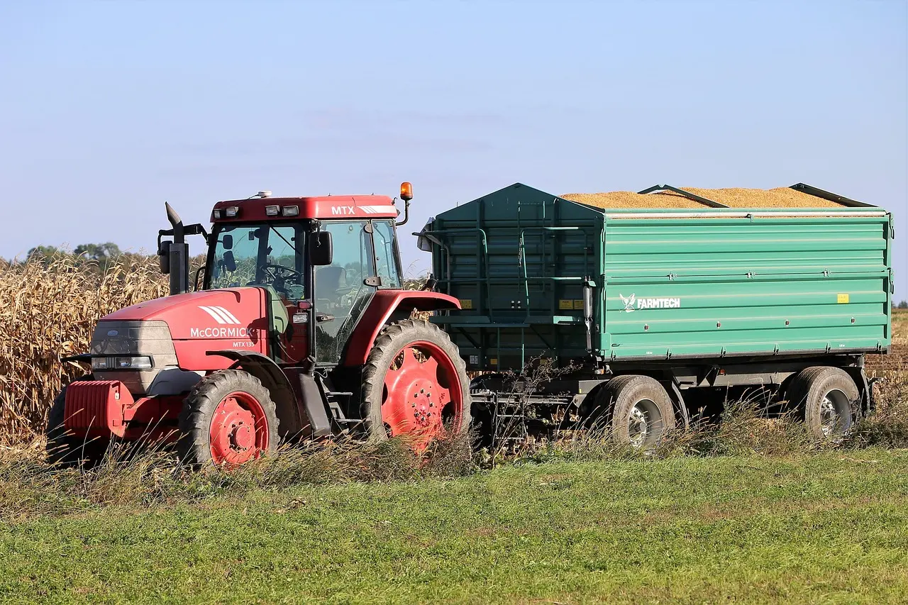 A vibrant red McCormick MTX tractor, hitched to a green FARMTECH trailer loaded with golden-brown harvested grain, sits in a vast field of tall, dry cornstalks. The clear blue sky highlights the scene, illustrating a key moment of transport in the agricultural harvest process.