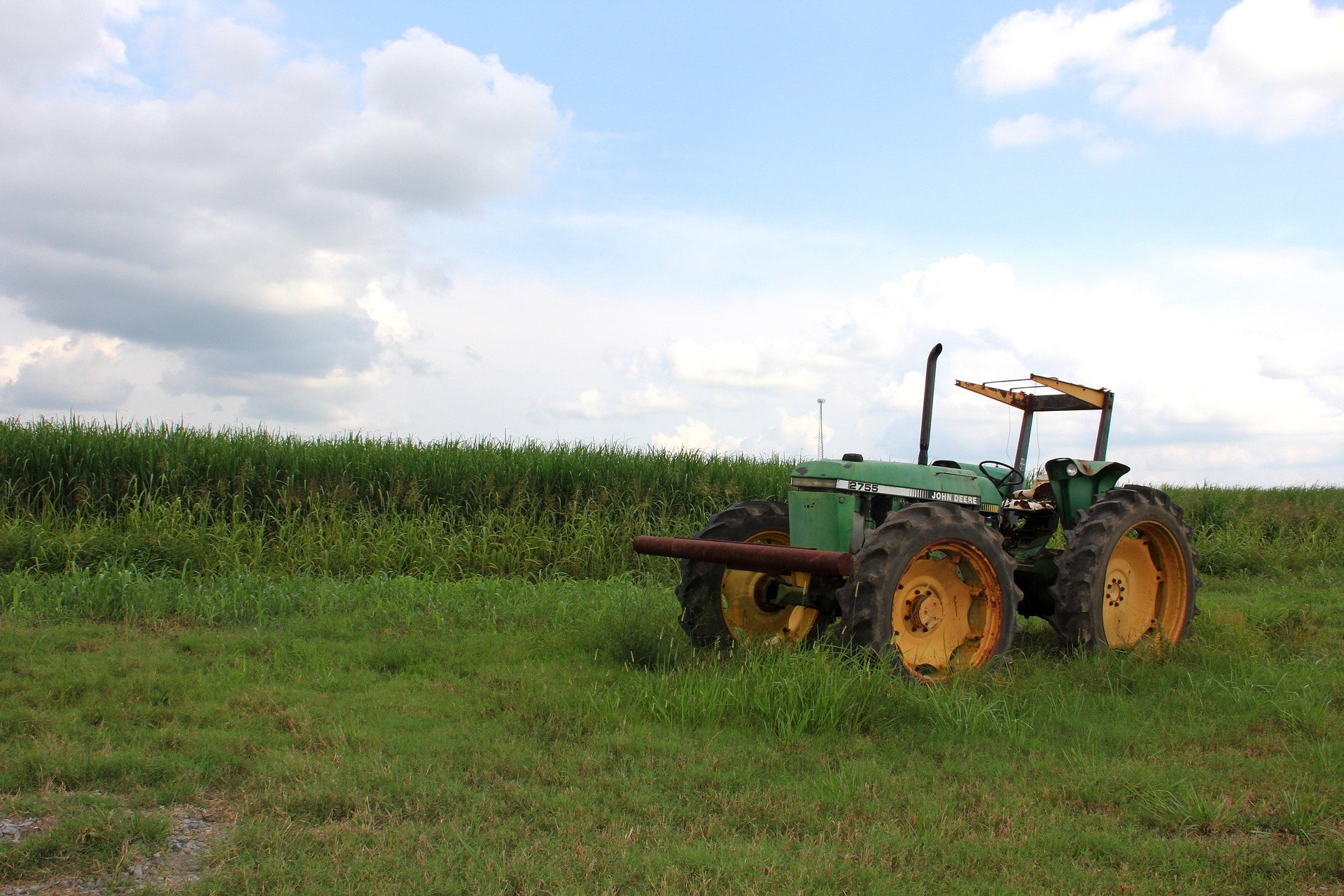 A green and yellow John Deere 2755 tractor is parked on a grassy verge, showcasing its classic design. The scene is set against a backdrop of a lush, tall cornfield under a partly cloudy blue sky, representing a quiet moment in the agricultural landscape.