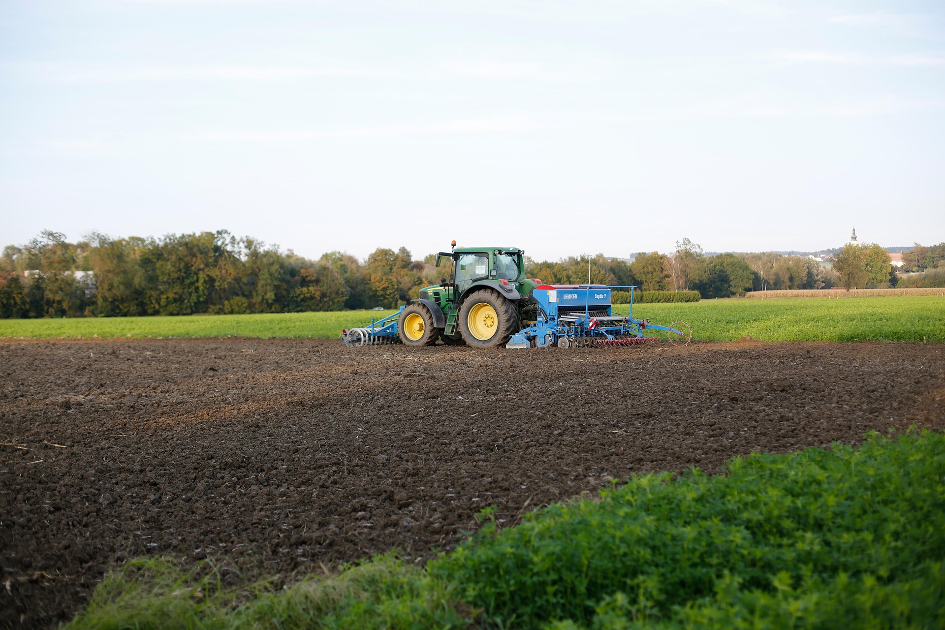 A green John Deere tractor actively pulls a blue Lemken Diamant seed drill across a field of dark, cloddy soil, laying precise parallel furrows for planting. The scene, with vibrant green vegetation in the foreground and a distant treeline under a soft sky, captures the precision and harmony of modern mechanized sowing.