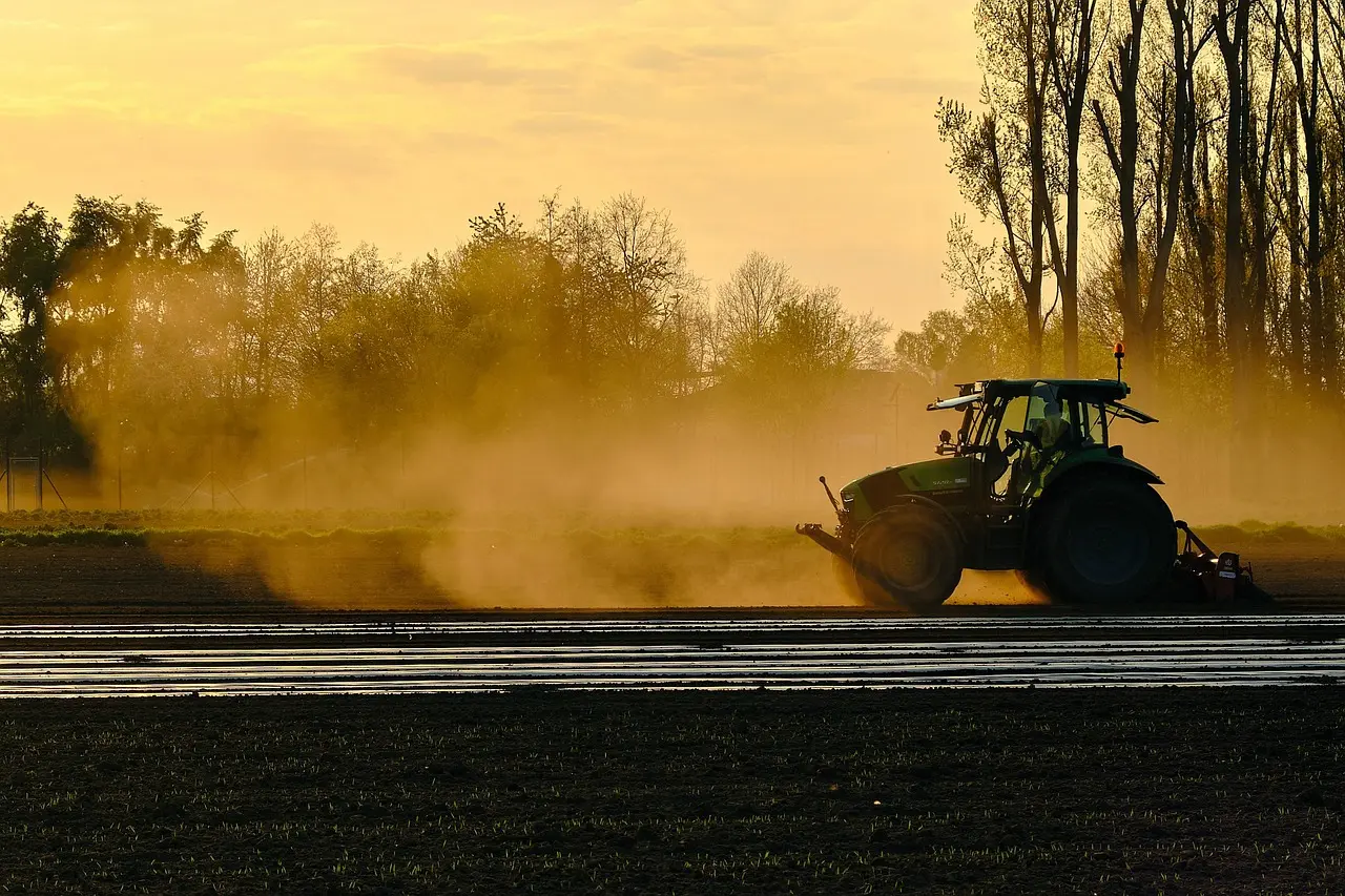 At golden hour, a dark green tractor creates a dramatic cloud of dust while tilling a field. The scene is deeply atmospheric, with reflective wet furrows in the foreground contrasting against the soft haze and the silhouette of trees under a vibrant orange-yellow sky, capturing the raw, poetic essence of mechanical farm labor at day's end.