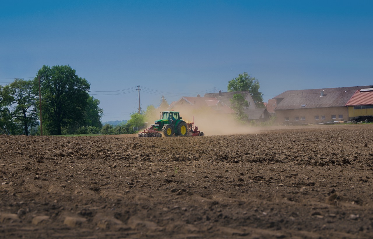 A green tractor with attached red machinery tills a brown field, creating a dynamic plume of dust. Set against a clear blue sky, the rural scene includes distant green trees and houses with yellow and brown roofs, illustrating the active progress of agricultural work.