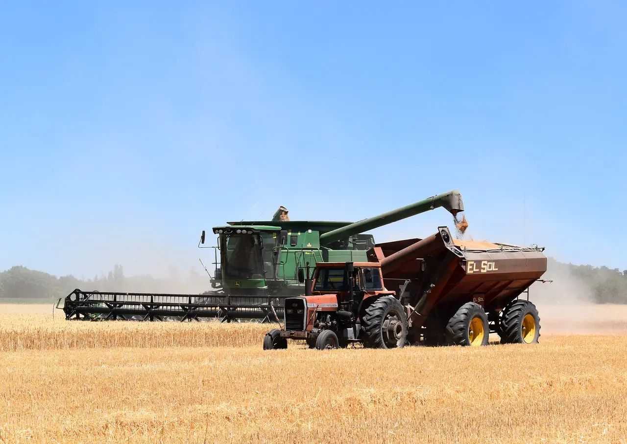 A green combine harvester transfers golden wheat into the "EL SOL" grain cart towed by a red tractor, showcasing efficient teamwork during harvest in a sunlit field under a cloudless blue sky.