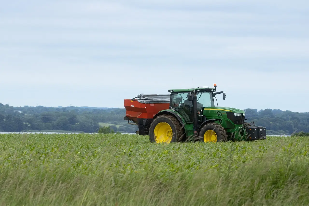 A green and yellow tractor transports a covered load on a red trailer across a serene, expansive field of young green crops, with rolling hills in the background under a mild sky, embodying orderly agricultural work.