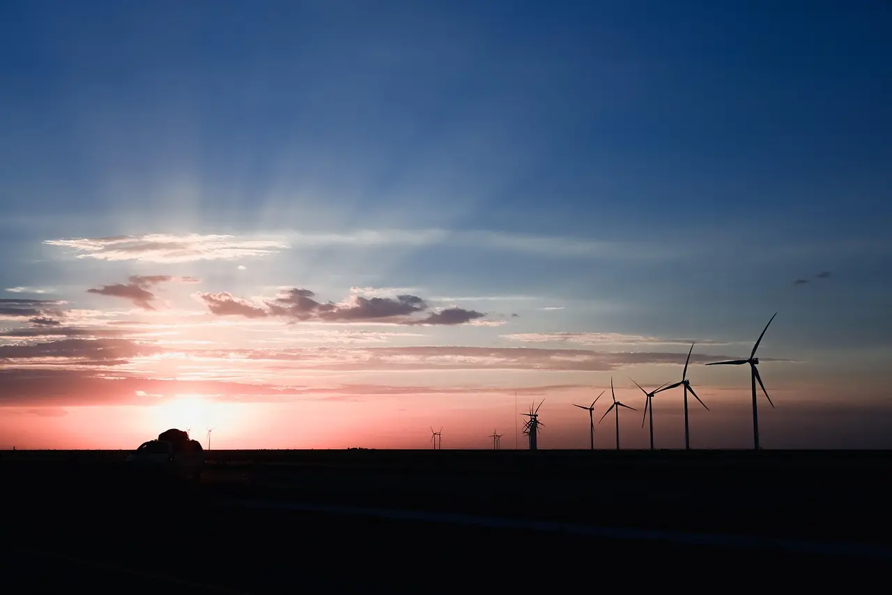 A warm, serene sunset scene features the soft glow of the sun behind clouds, casting a gradient of orange and red into a deep blue sky. Silhouettes of wind turbines stretch along the horizon, symbolizing sustainable energy, with a car in the foreground suggesting a moment of pause on a journey, evoking a sense of peace and transition.