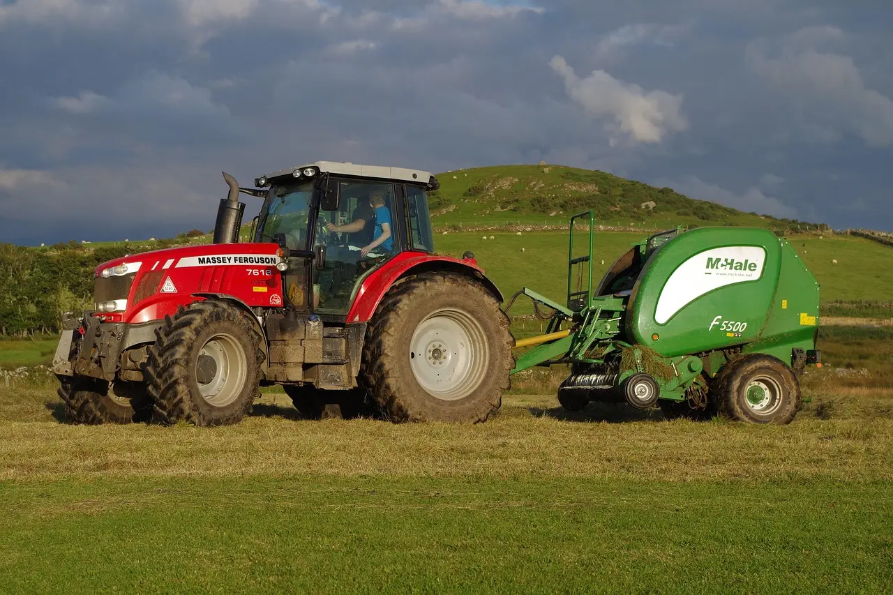 A red Massey Ferguson 7618 tractor, with a visible operator in the cab, tows a green M-Hale F5500 forage wagon across a neatly maintained pasture. Set against a backdrop of rolling green hills dotted with grazing sheep under a cloudy sky, the scene exemplifies the integration of precise machinery with traditional pastoral farming.