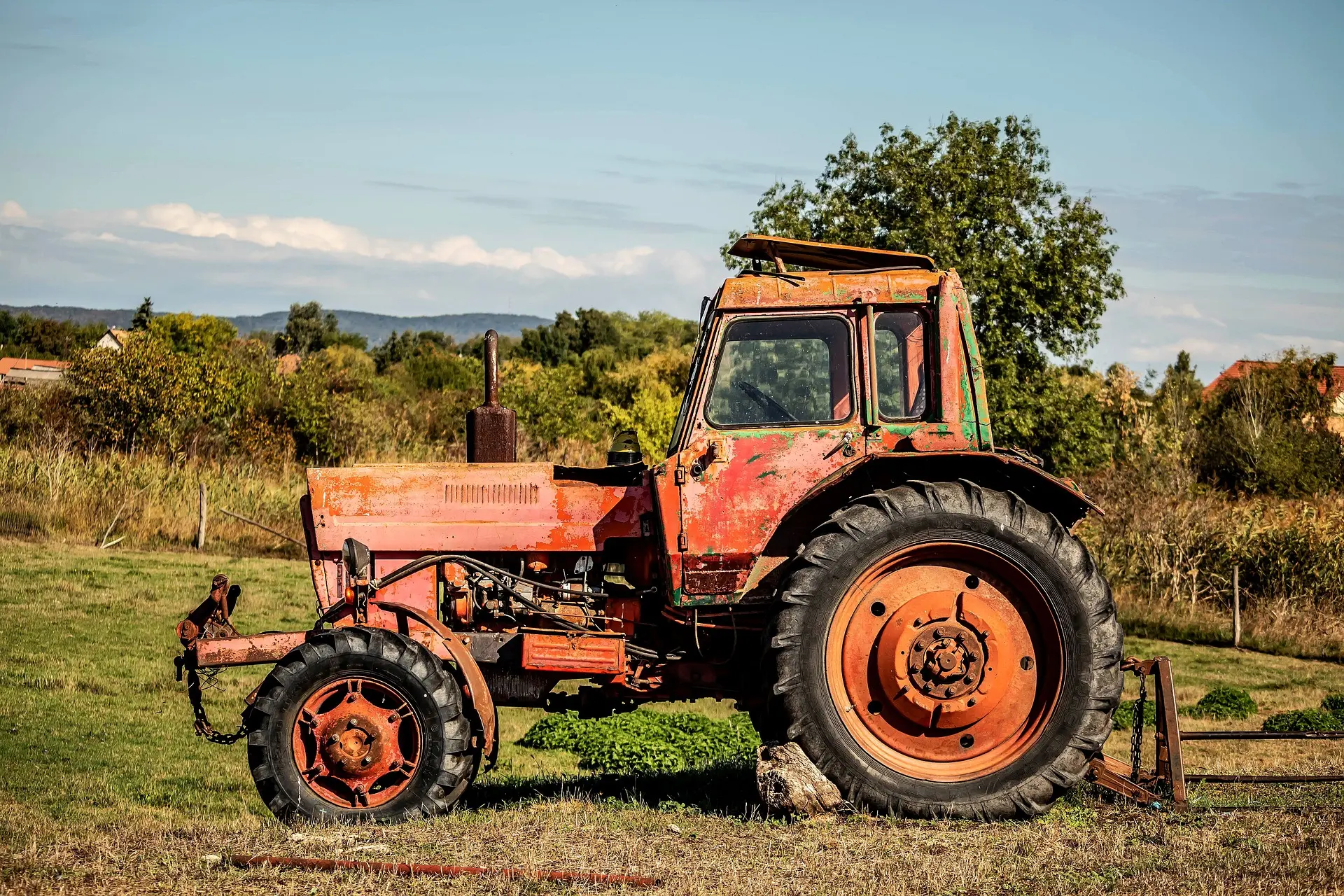 An old, heavily rusted orange tractor with extensively peeling paint rests silently in a field of golden grass, enclosed by a rustic wire fence. Set against a backdrop of lush trees and a soft blue sky with wispy clouds, the image evokes a profound sense of nostalgia, stillness, and the passage of time in the countryside.