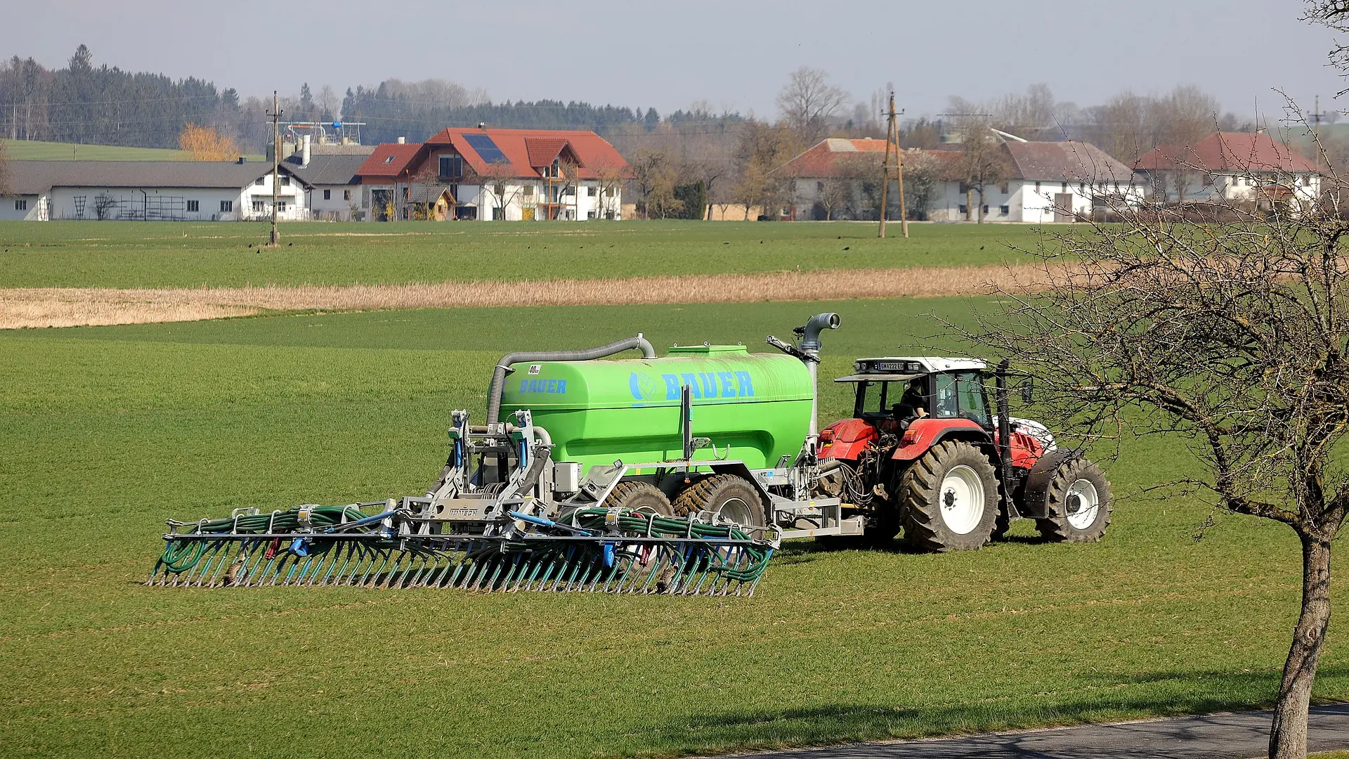 A red agricultural tractor is connected to a sizable green "BAUER" storage tank or applicator, positioned in a lush green field. Set against a backdrop of rural houses with red and gray roofs amidst trees, the scene illustrates the integration of farm machinery and infrastructure within a peaceful countryside setting.