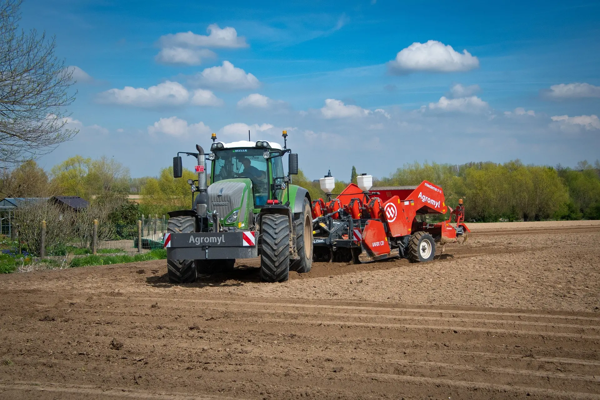 A green Agromyl-branded tractor, with the model name “VARICO 38” visible, pulls a red Agromyl agricultural implement across a tilled field. The scene, set under a clear blue sky with scattered clouds and green trees in the distance, clearly showcases the machinery in action and represents modern, mechanized farming operations.