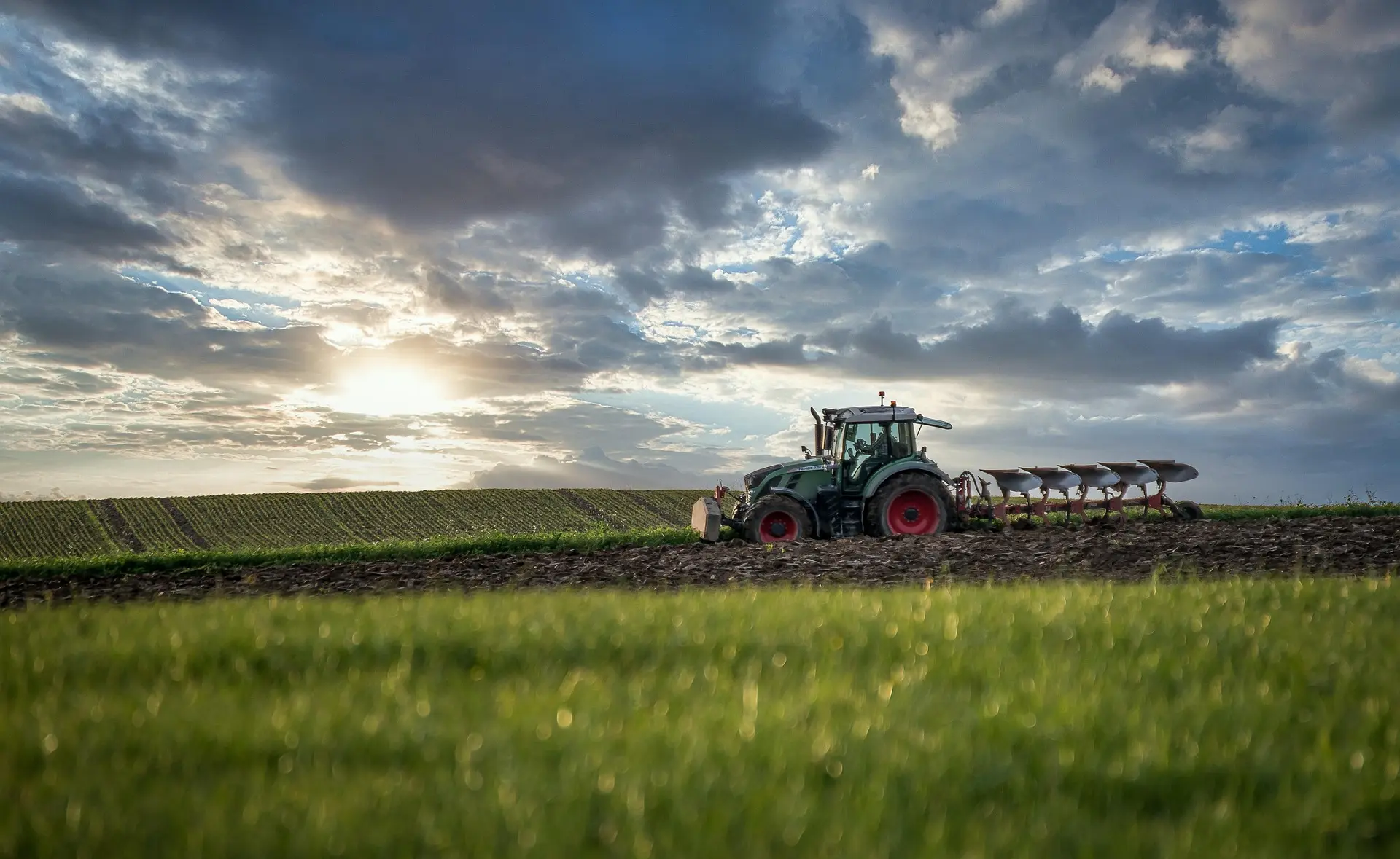 A green tractor with bright red tires works across a field, connected to an implement. The scene is set against a backdrop of orderly, layered fields, under a dynamic sky where sunlight breaks through heavy clouds, casting dramatic light and shadow across the peaceful, productive rural landscape.