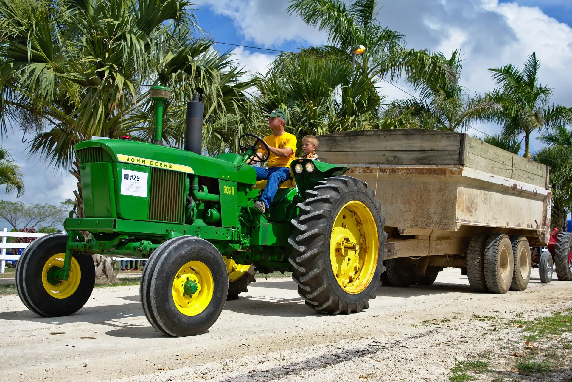 A man in a yellow shirt and a boy ride together in the cab of a vibrant green and yellow vintage John Deere 3020 tractor, towing a sizable wooden trailer down a white gravel road. Lush palm trees line the path under a bright, sunny sky with scattered clouds, creating a warm scene that blends family, legacy, and agricultural work.