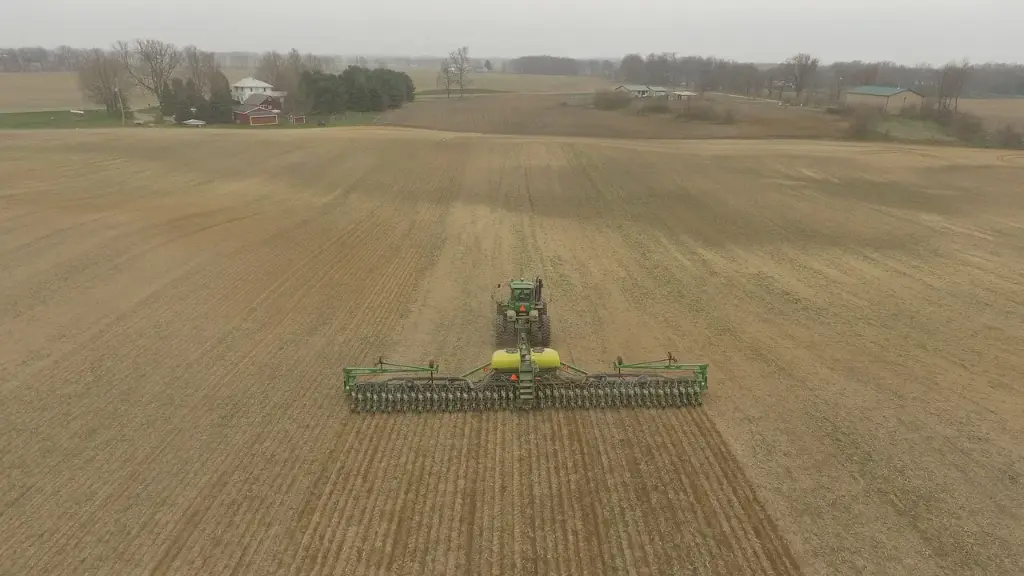 An aerial photograph captures a green tractor and its implement creating precise furrows in a vast, open brown field. The orderly patterns of tilled soil dominate the scene, which is set against a soft grey sky with distant farmhouses featuring red and white roofs nestled among trees, conveying the scale and rhythm of field work.