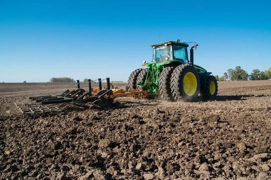 A green John Deere tractor, prominently displaying its brand logo, actively pulls a bright yellow agricultural implement through rich brown soil. The scene is set under a clear blue sky with a distant tree line, and features the text “Don Cachão,” highlighting a moment of productive fieldwork with modern machinery.