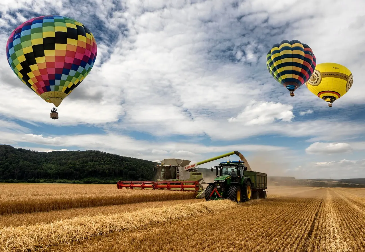 A green tractor with the letter “D” on its side pulls a red combine harvester during harvest in a golden wheat field. The scene blends modern agriculture with leisure, featuring vibrant hot air balloons—one multicolored and two striped—drifting over lush hills under a partly cloudy sky.