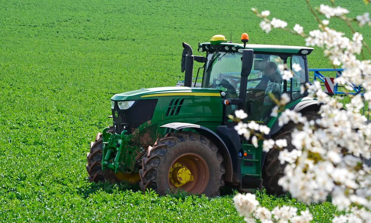 A powerful, dark green tractor with a closed cab and prominent yellow/orange warning lights is positioned in a lush, green field. An operator is visible through the cab window. In the softly blurred foreground, branches of white flowers add a touch of spring, highlighting the active yet harmonious scene of modern machinery at work in nature.