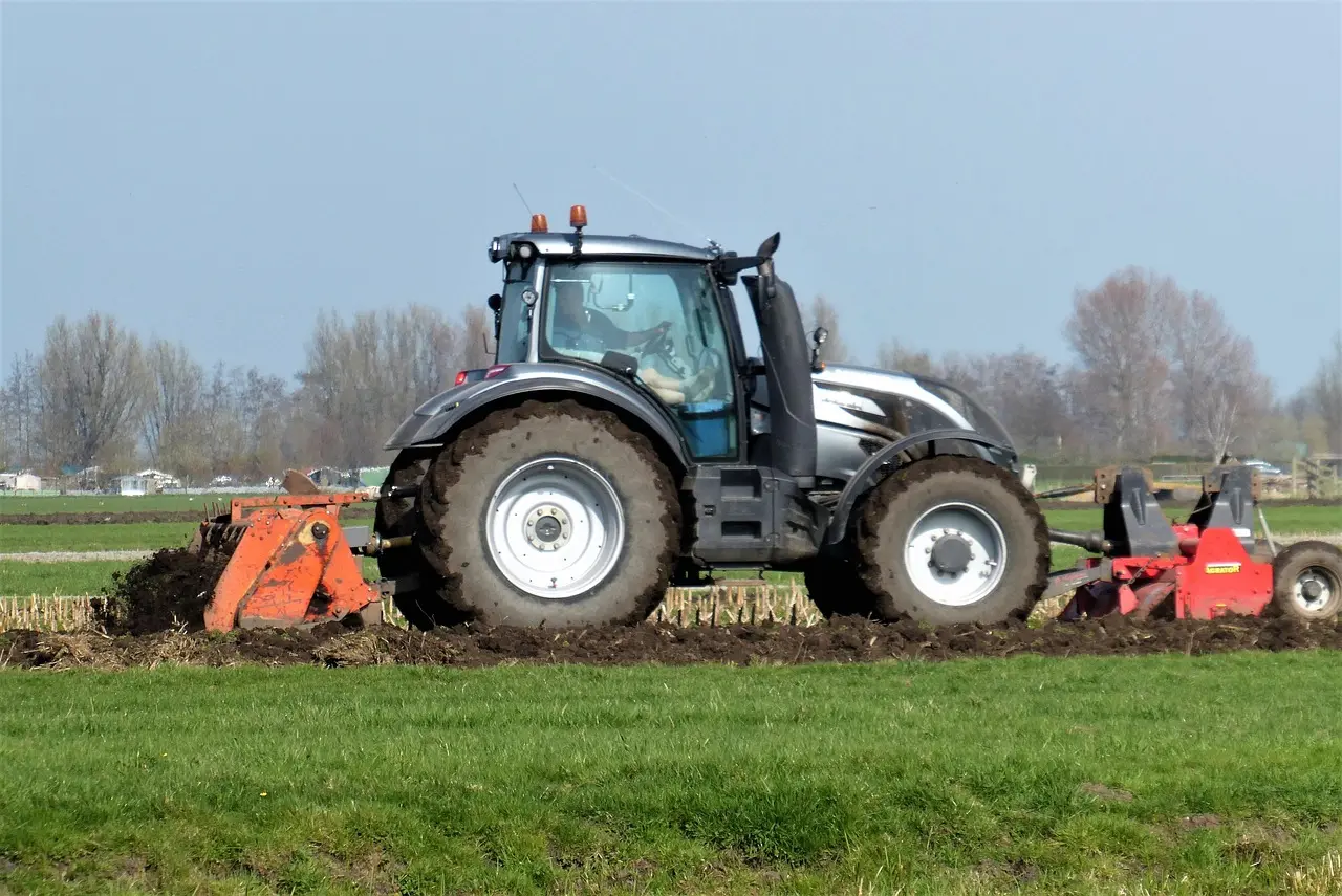 A robust, silver-gray tractor, its wide tires covered in soil and equipped with prominent orange safety lights, actively pulls a red tillage tool across a vast green field. Set against a backdrop of distant trees and farmhouses under a light blue sky, this image captures the essence of modern, mechanized agriculture in a serene landscape.