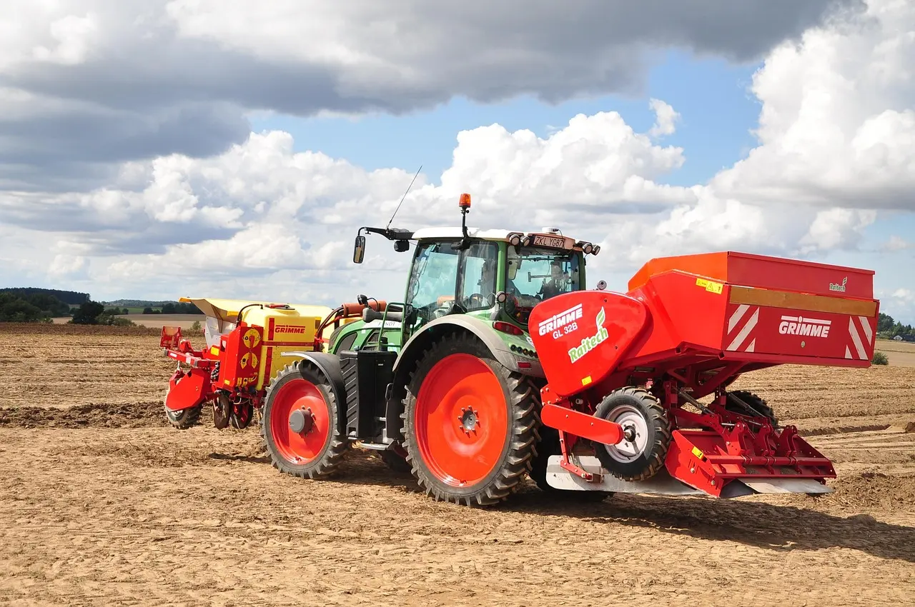 A modern GRIMME tractor in green and black, with an orange beacon and red wheel hubs, operates in a field. It is attached to a large red Rotech GL 32B implement, with another yellow GRIMME machine beside it. This image showcases the precise application of modern agricultural technology in an open field under a cloudy sky.
