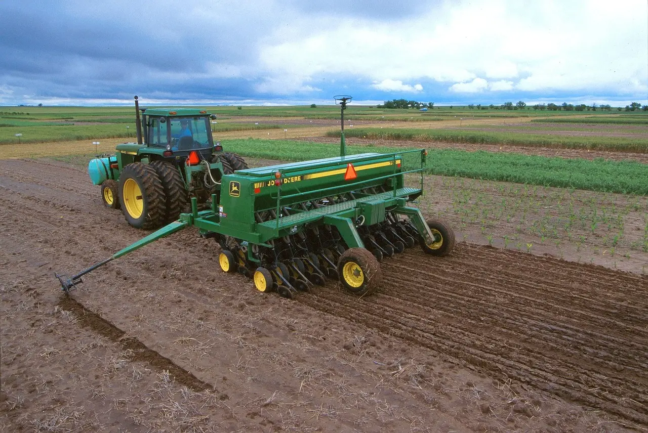 A green John Deere tractor, with prominent yellow wheel rims and a visible operator in the cab, pulls a green John Deere planter/fertilizer applicator across a vast, tilled brown field. The contrasting left side with fresh furrows and the right side with established green crop rows illustrates the process of precision planting under a heavy, cloudy sky, showcasing modern mechanized agriculture at work.