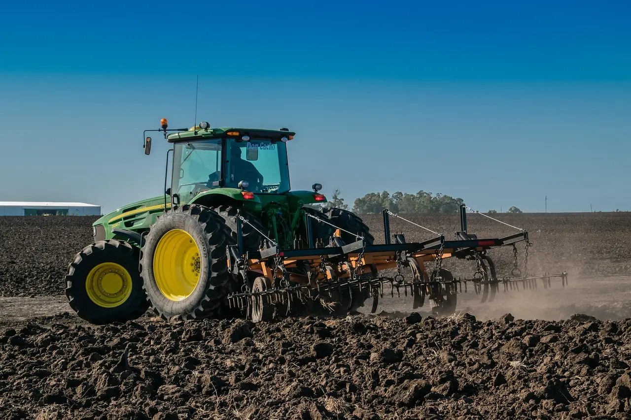 A powerful green and yellow John Deere tractor plows a field, its large rear tires and attached implement churning the earth and creating a dynamic dust cloud. Set against a vast open field and clear blue sky, this image captures the strength and motion of modern mechanized farming in a bright, productive landscape.