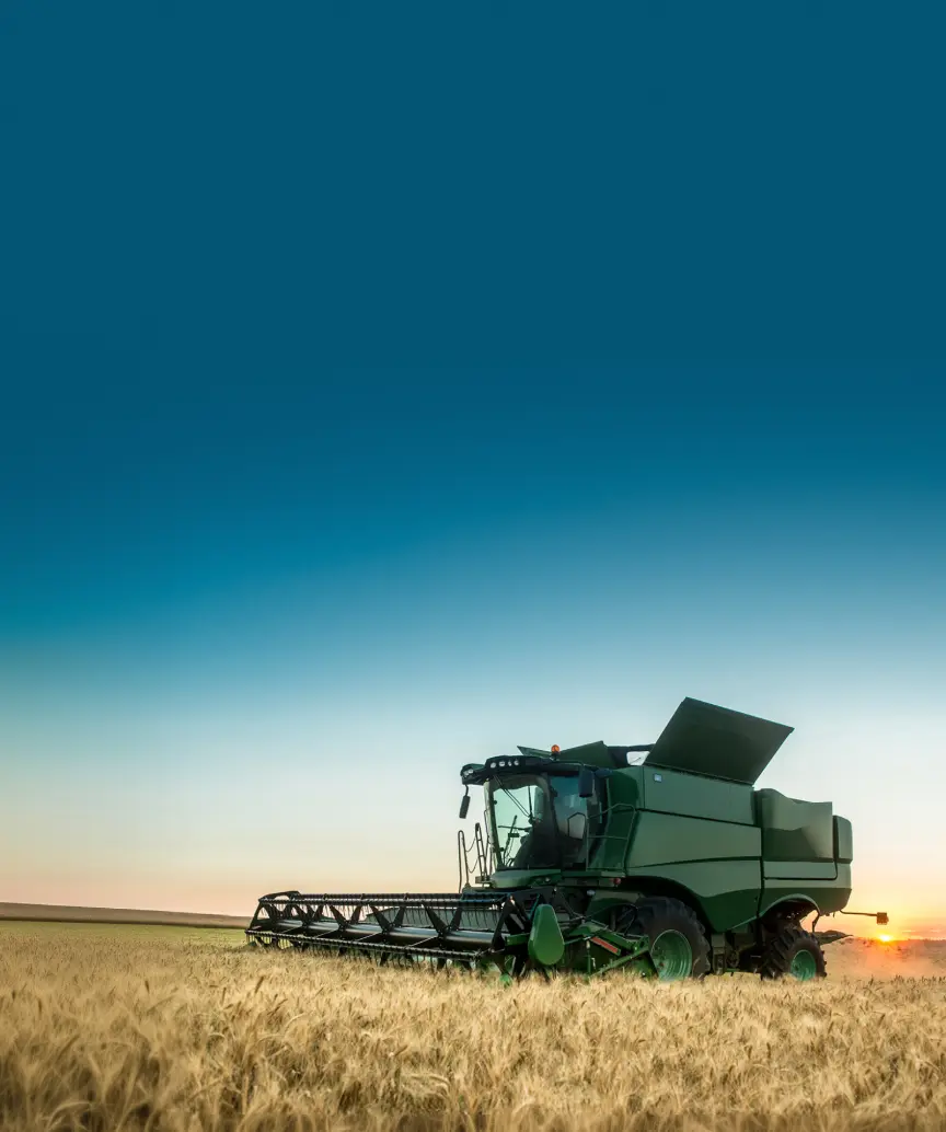 A green combine harvester works in a golden wheat field under a dramatic sunrise or sunset sky. With its header cutting and unloading arm raised, the machine is silhouetted against vibrant gradients of orange, yellow, and blue, symbolizing the efficiency and beauty of modern harvest.