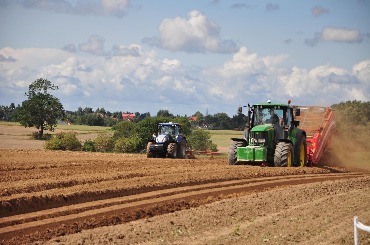 Multiple tractors at work in a vibrant rural landscape. In the foreground, a green tractor actively pulls a red tillage implement through dark brown, neatly ridged soil, creating a plume of dust. Another blue tractor operates in the mid-ground, with green trees, houses, and distinctive red-roofed buildings completing the scene under a partly cloudy sky, evoking a sense of productive energy in the countryside.