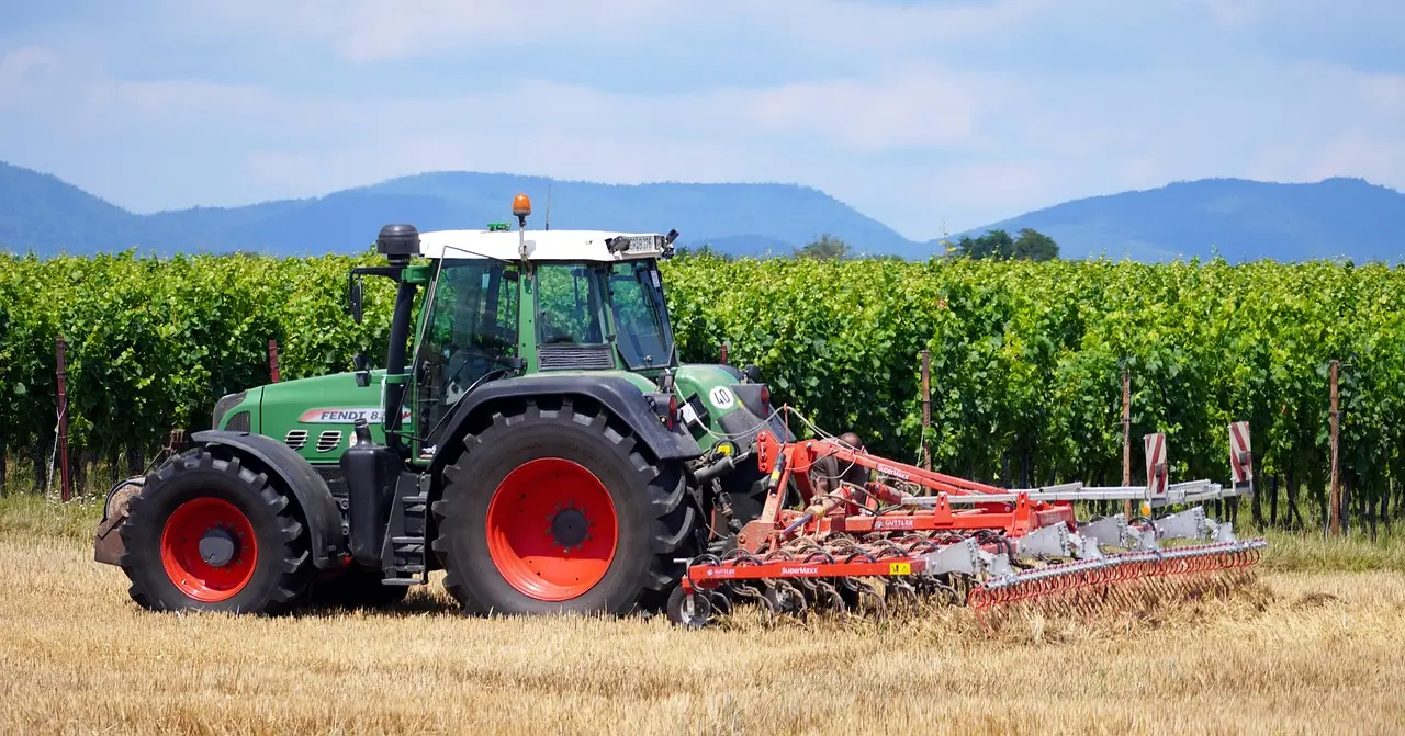 A powerful green Fendt 800 series tractor, featuring large tires with bright red rims and an orange roof beacon, is parked in a harvested field while hitched to a red agricultural implement. The serene scene is set against a backdrop of orderly, lush green vineyard rows and soft-focus distant mountains under a clear sky with clouds, showcasing modern farm machinery within a peaceful pastoral setting.