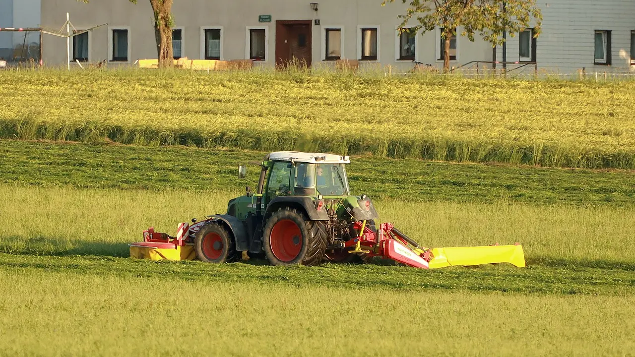 A large green Fendt tractor operating a red and yellow agricultural implement in a sunlit, freshly cut green field during late afternoon, with a building in the background.