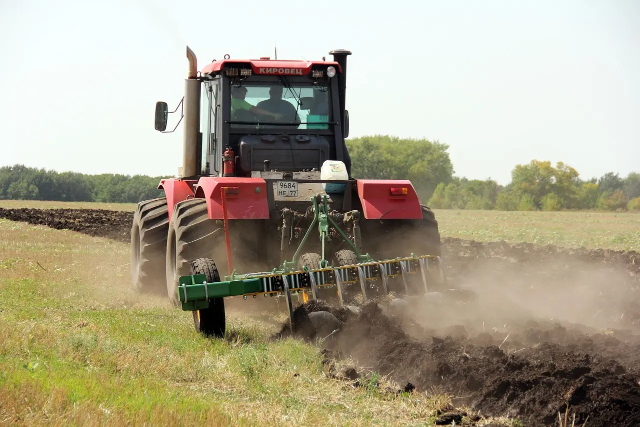 A powerful red Kirovets tractor (license plate 9684 HE 77) vigorously cultivates soil, kicking up a dust cloud, with a green implement in a vast field under a blue sky.