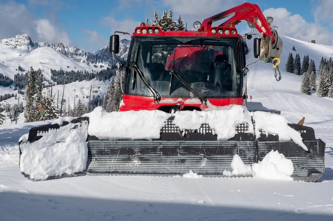 A red pistenbully snow grooming vehicle with a large black snowplow blade pushes and smooths fresh white snow on a ski slope, with snow-covered mountains and trees under a clear blue sky, highlighting essential winter sports area maintenance.