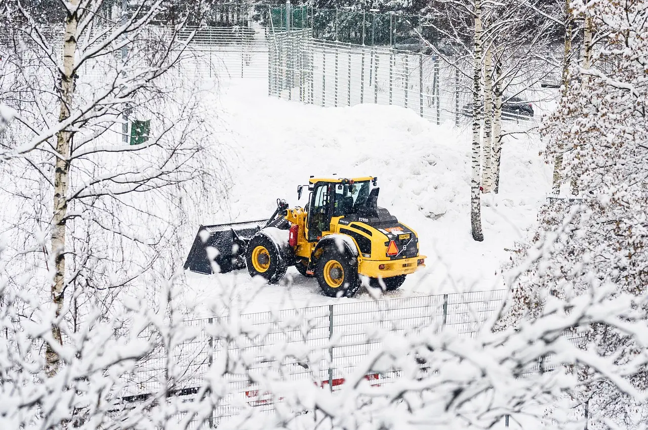 A yellow and black bulldozer sits stationary in a deep snowscape, its bucket piled with snow, surrounded by snow-laden trees and metal fencing near resort buildings, depicting a pause during winter clearing operations.