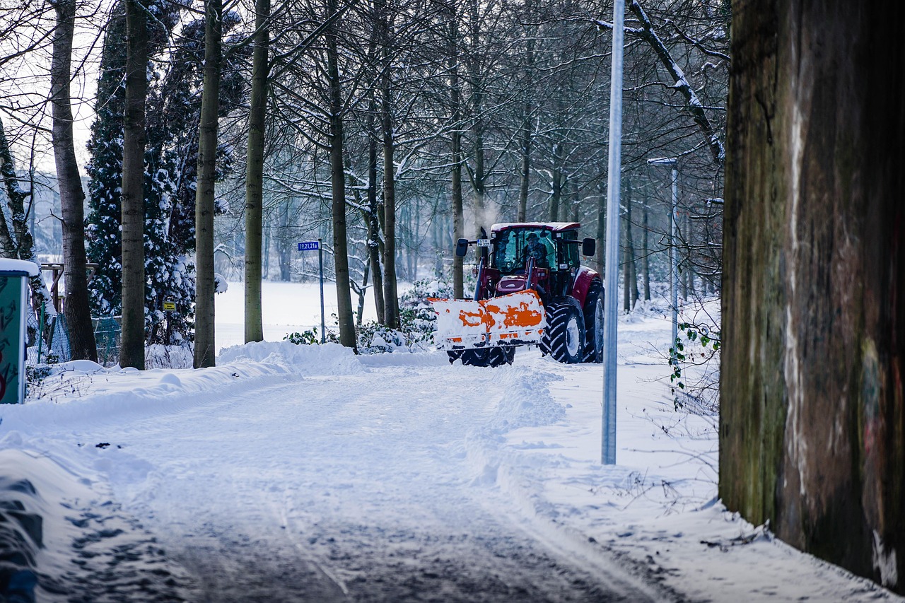 A red tractor with an orange snowplow attachment clears a snow-covered road during a winter maintenance operation, flanked by trees, shrubs, and a stone wall under a soft gray sky.