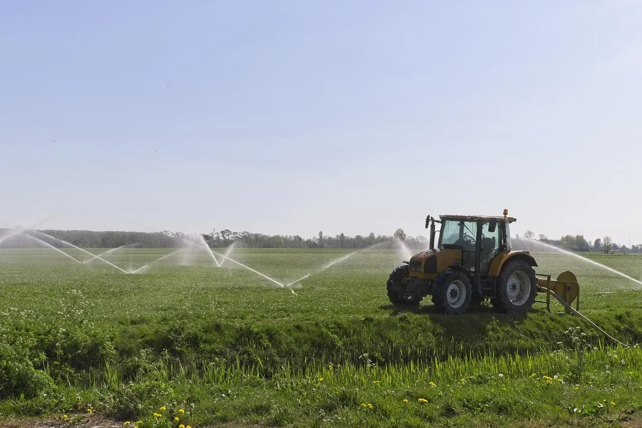 A yellow tractor pulls a mobile irrigation system across a farm field, with multiple sprinklers emitting fan-shaped water sprays to irrigate lush green crops dotted with yellow flowers, under a light blue sky with distant trees.