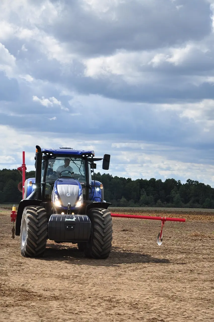 A large blue and black tractor tows a red agricultural implement (such as a planter or cultivator) while working in a vast, light-brown field. A driver is visible inside the cab, and the tractor's headlights are on. The background features dark green trees and a cloudy sky.