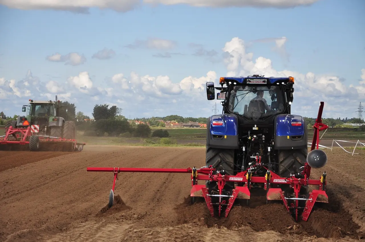 Two tractors work together in a field. In the foreground, a blue tractor with the "GRIMME" logo pulls a red tillage implement, kicking up dust. Another darker-colored tractor operates farther away. The scene includes distant trees, vegetation, house outlines, and a clear sky with clouds.