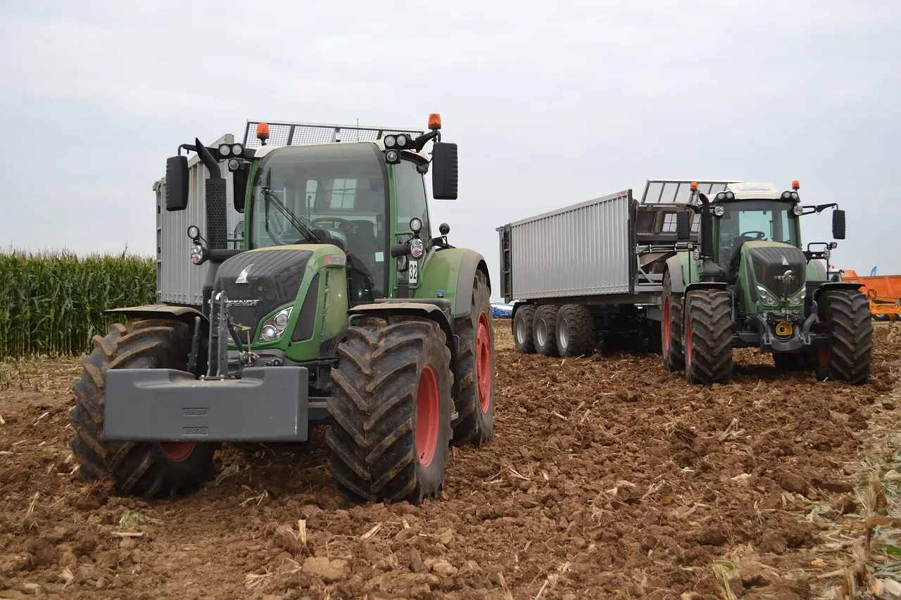 Two large, green John Deere tractors (marked with the number "32" on one) transport materials across freshly tilled farmland. The lead tractor has wide black tires, multiple orange beacon lights, and a front ballast weight, while the following tractor pulls a long, sturdy silver trailer with multiple wheels. A row of green cornfields lines the background under a cloudy sky, showcasing modern agricultural logistics.